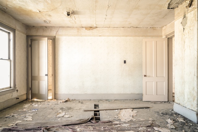 A white room with peeling paint, missing floorboards, and which is in a state of disrepair.